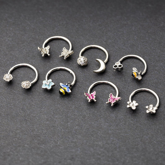 Silver horseshoe rings featuring various charms, including butterflies, bees, moons, and flowers, displayed on a dark background.
