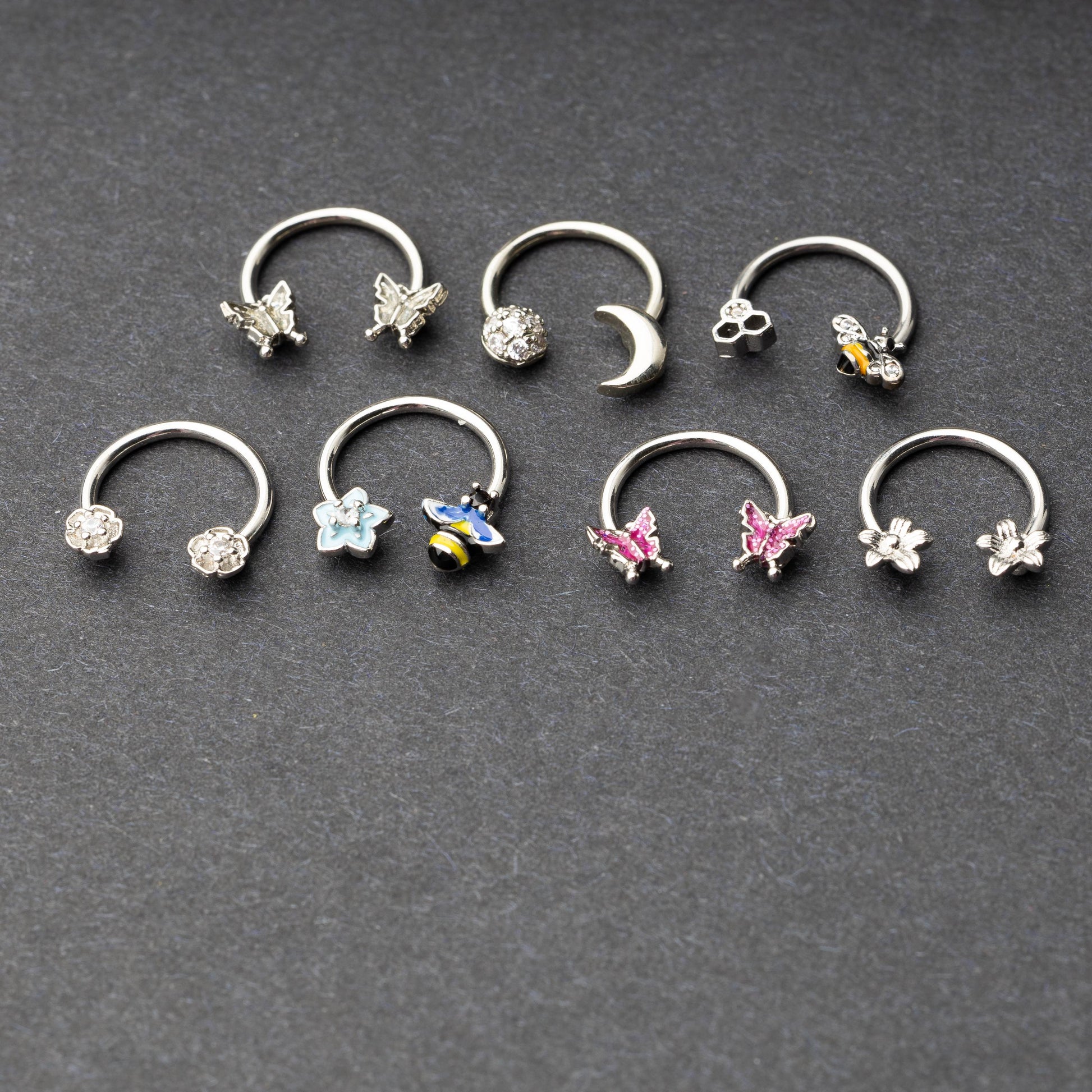 Silver horseshoe rings featuring various charms, including butterflies, bees, moons, and flowers, displayed on a dark background.