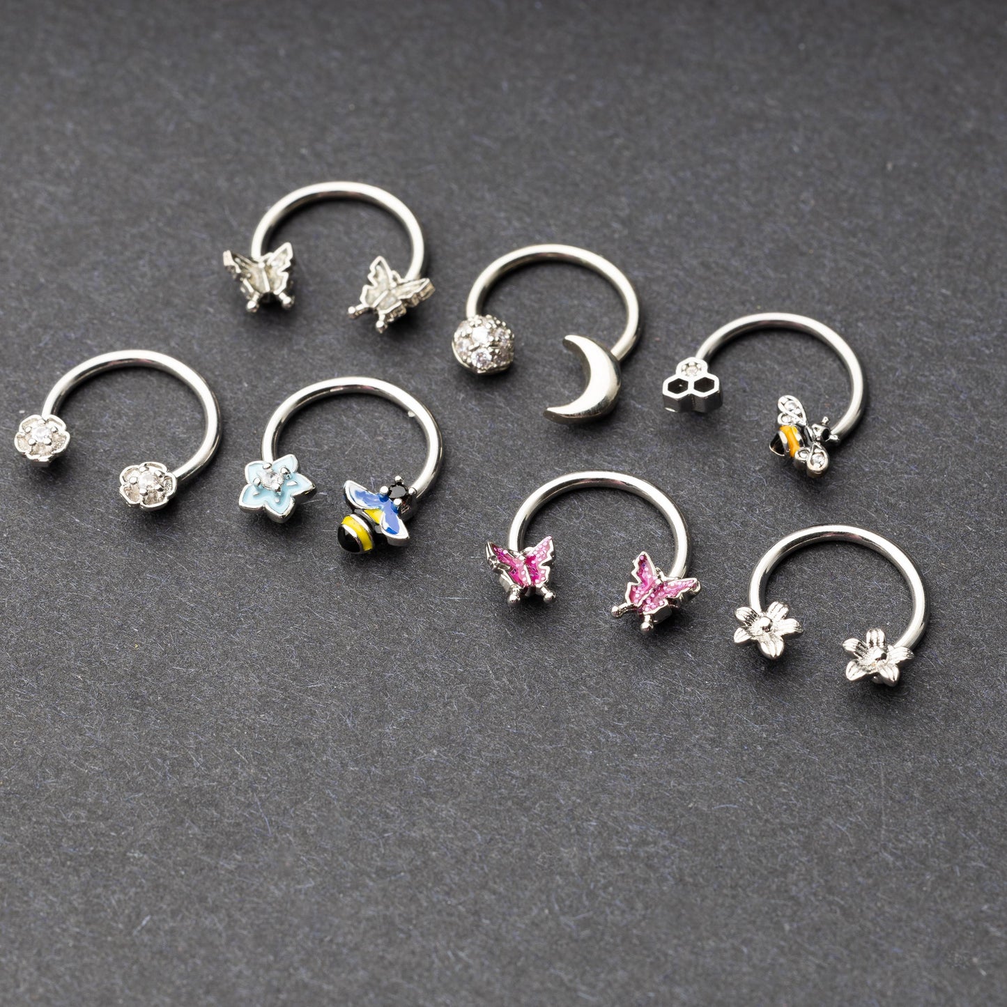 Silver horseshoe rings featuring various charms, including butterflies, bees, moons, and flowers, displayed on a dark background.