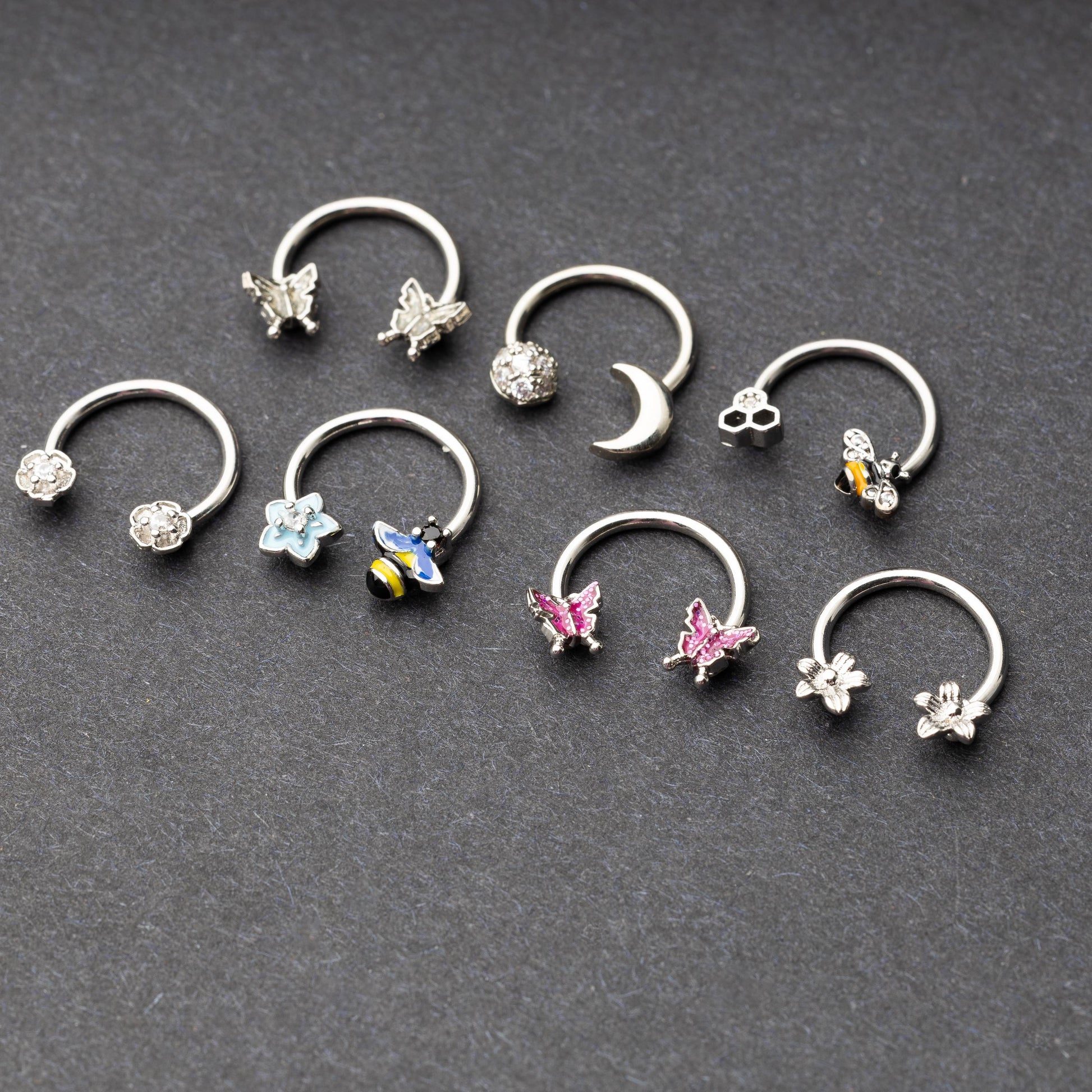 Silver horseshoe rings featuring various charms, including butterflies, bees, moons, and flowers, displayed on a dark background.