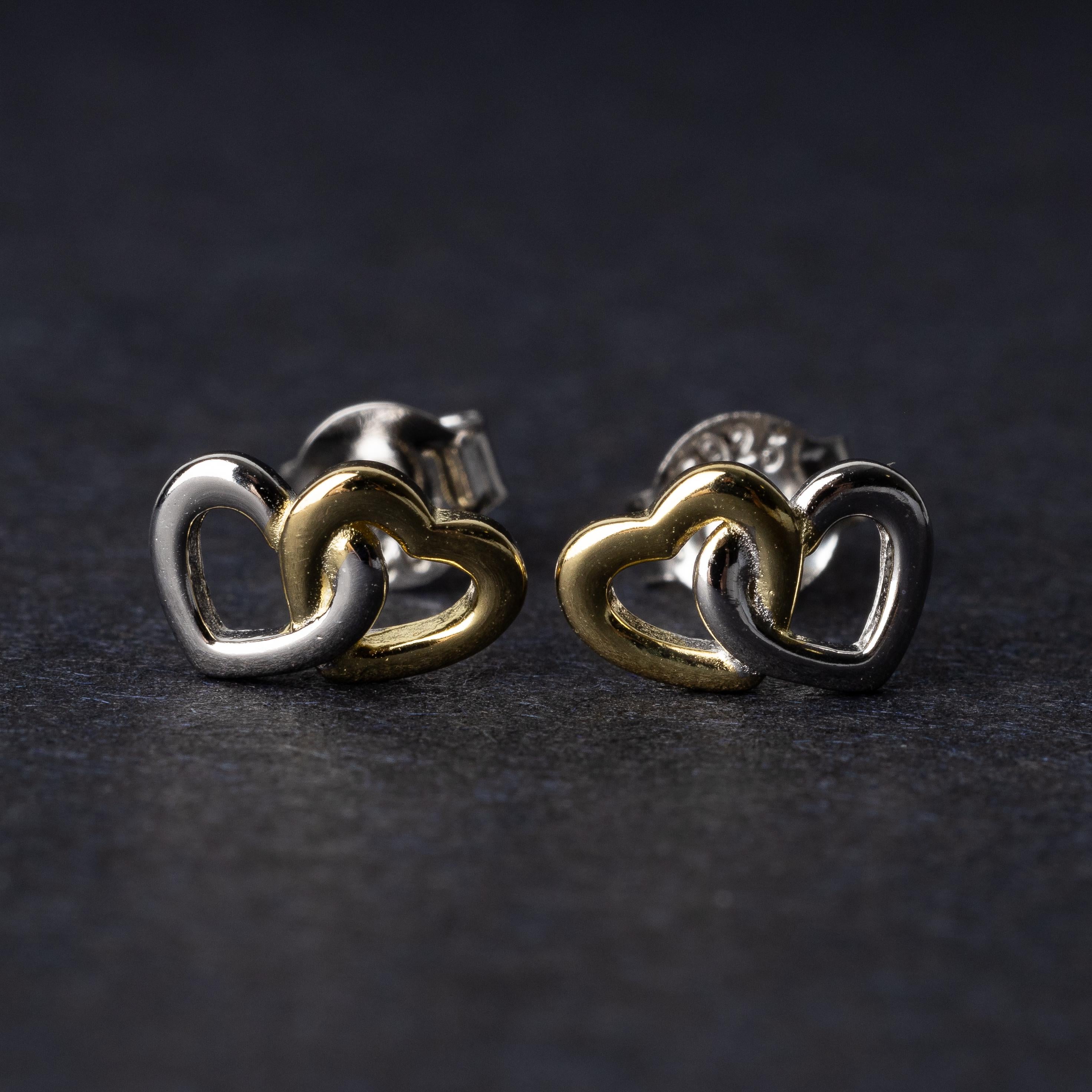 Close-up of interlocking silver and gold heart stud earrings with a polished finish, displayed on a dark background.
