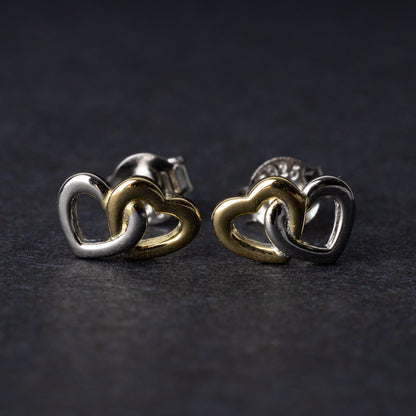 Close-up of interlocking silver and gold heart stud earrings with a polished finish, displayed on a dark background.