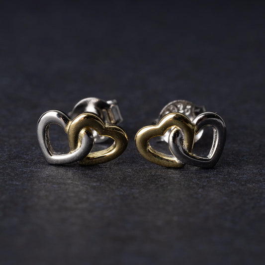 Close-up of interlocking silver and gold heart stud earrings with a polished finish, displayed on a dark background.