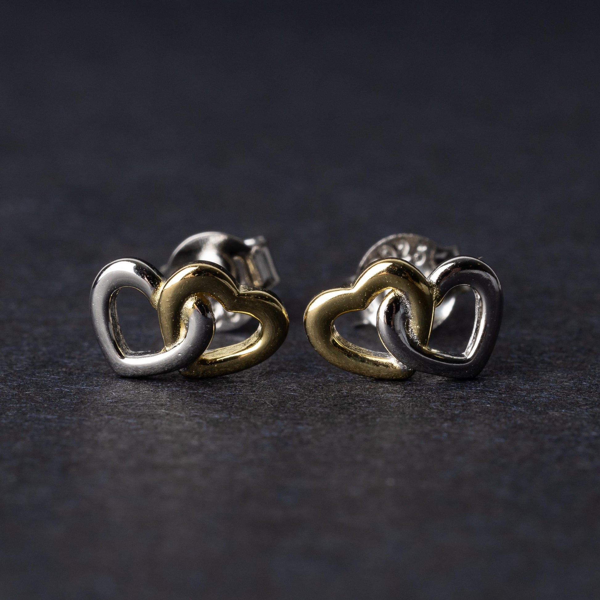 Close-up of interlocking silver and gold heart stud earrings with a polished finish, displayed on a dark background.