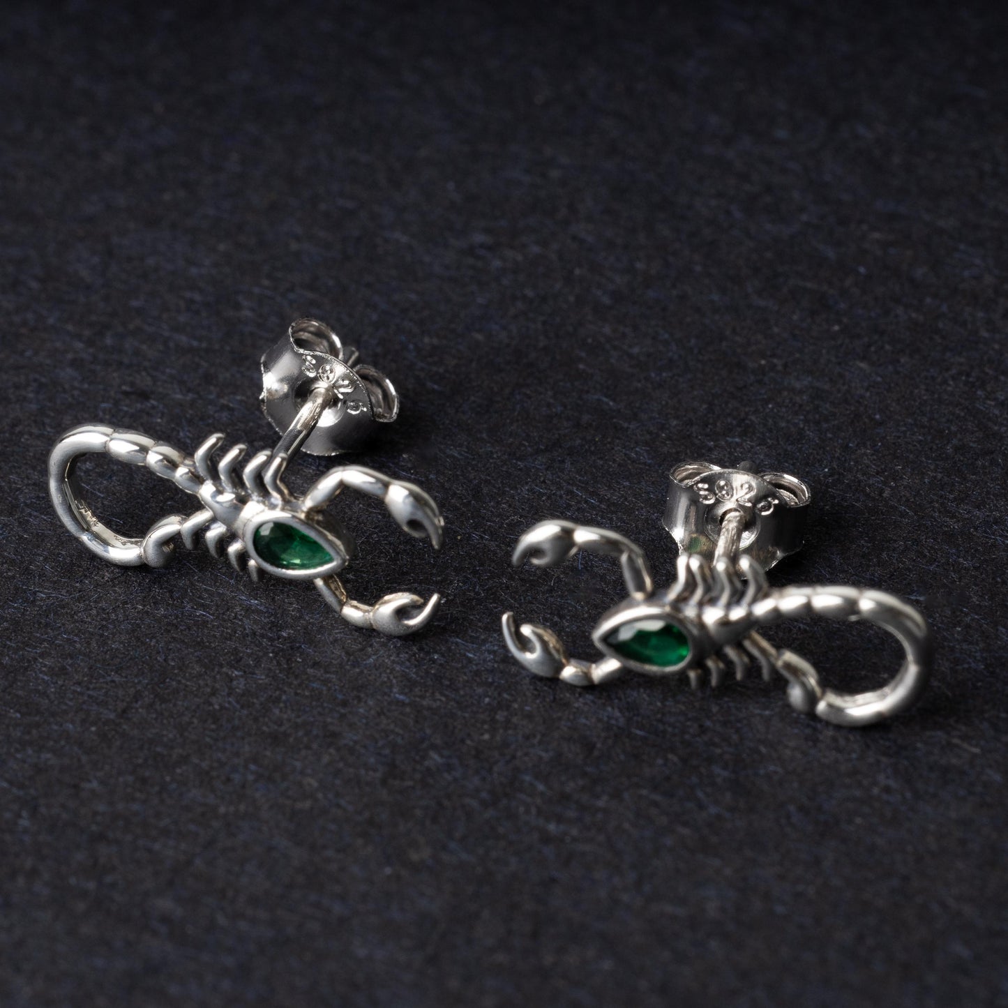 A pair of 925 sterling silver scorpion-shaped earrings with green gemstone accents as eyes, displayed on a dark background.