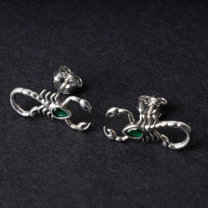 A pair of 925 sterling silver scorpion-shaped earrings with green gemstone accents as eyes, displayed on a dark background.