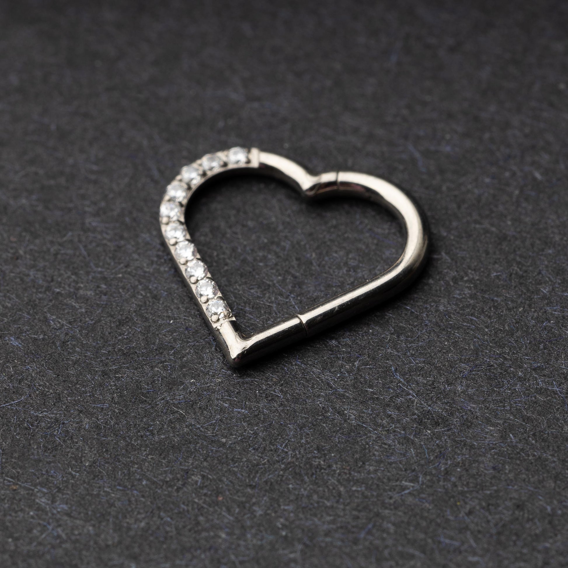 Heart-shaped daith clicker with crystal accents along one side, displayed on a dark textured background.