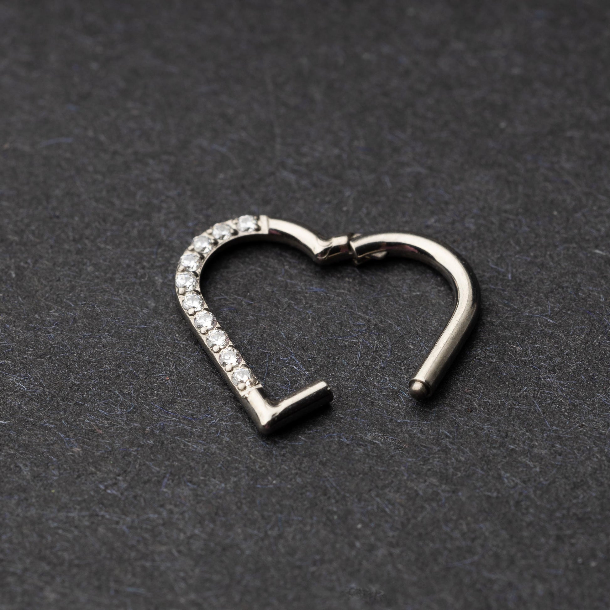 Heart-shaped daith clicker with crystal accents along one side, displayed on a dark textured background.