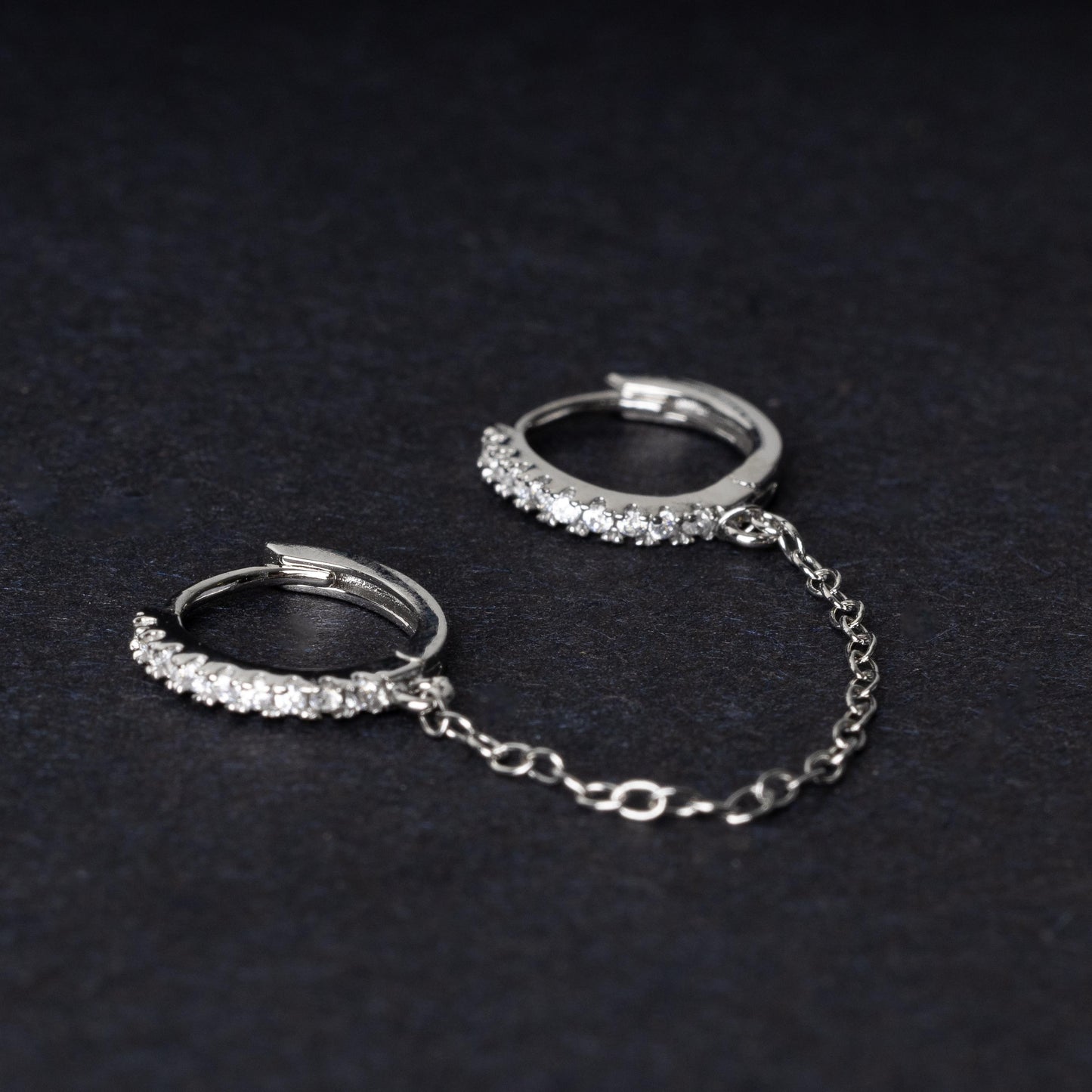 Silver huggie earrings with sparkling cubic zirconia detailing, connected by a delicate chain, displayed on a dark background.