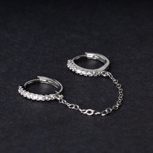 Silver huggie earrings with sparkling cubic zirconia detailing, connected by a delicate chain, displayed on a dark background.