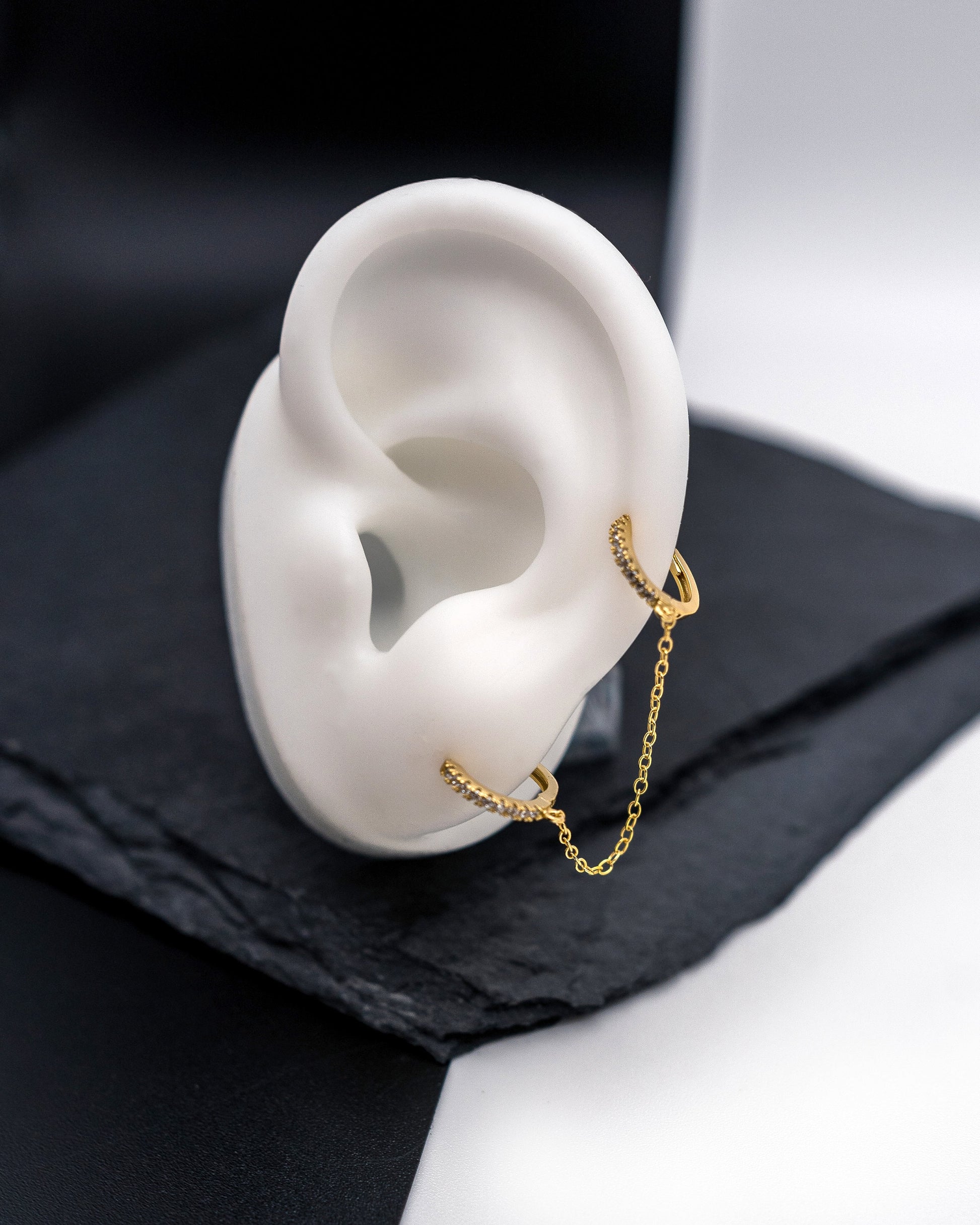 Gold huggie earrings with sparkling cubic zirconia detailing, connected by a delicate chain, displayed on a dark background.