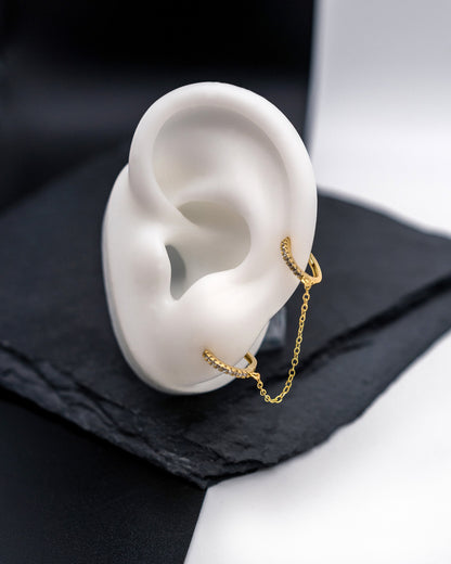 Gold huggie earrings with sparkling cubic zirconia detailing, connected by a delicate chain, displayed on a dark background.