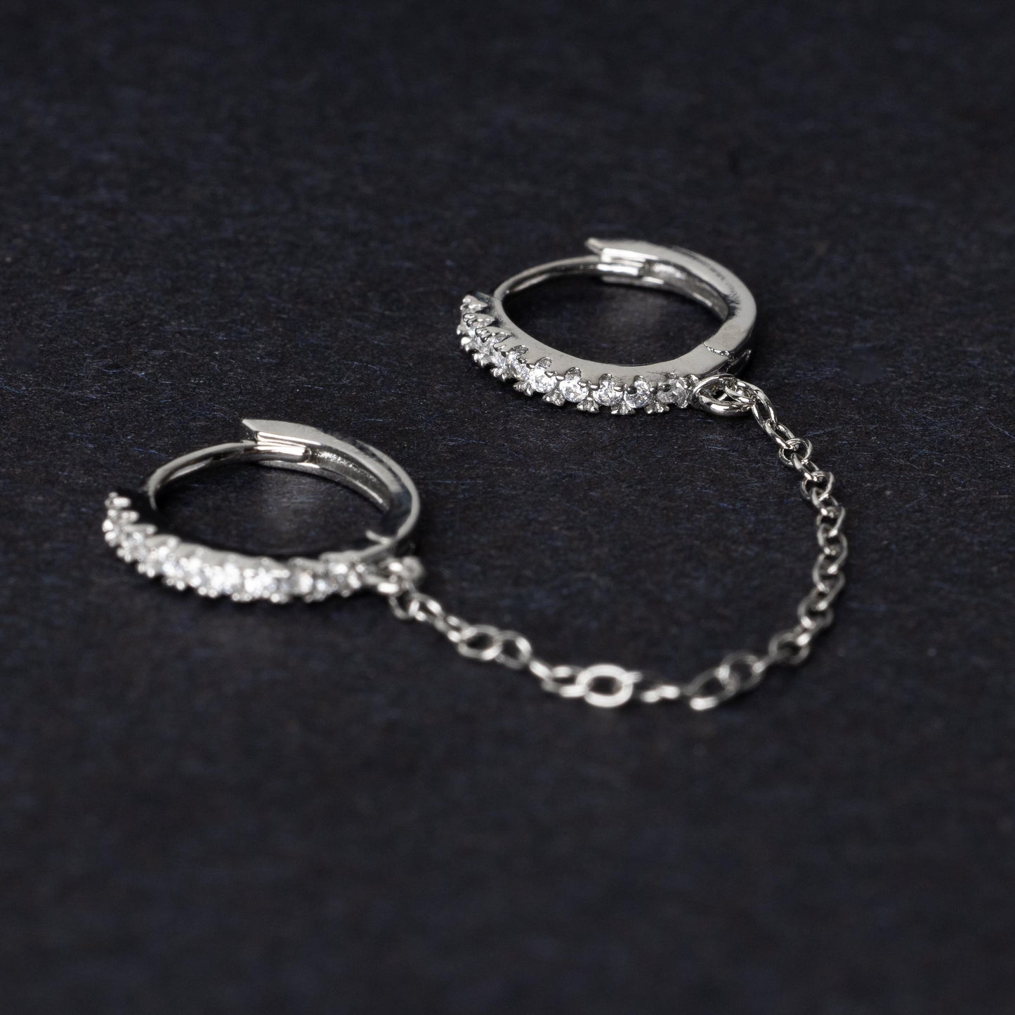 Silver huggie earrings with sparkling cubic zirconia detailing, connected by a delicate chain, displayed on a dark background.