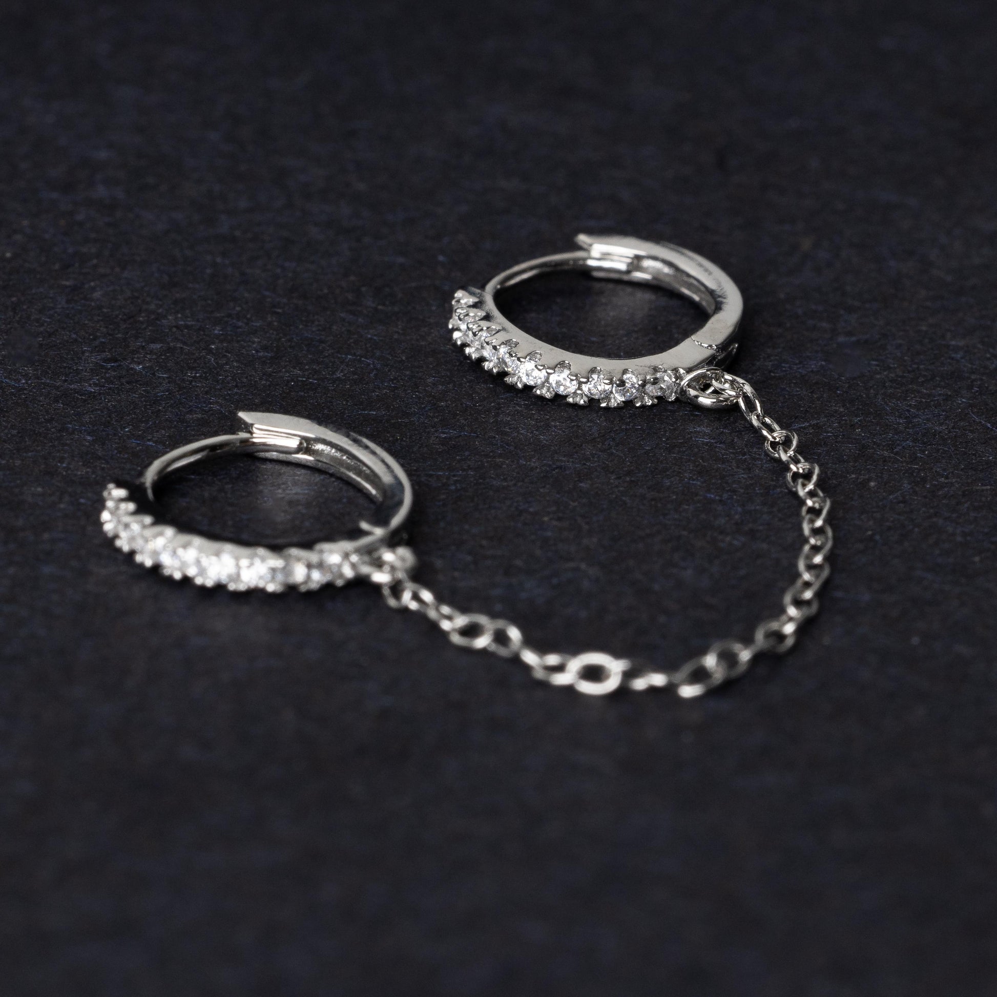 Silver huggie earrings with sparkling cubic zirconia detailing, connected by a delicate chain, displayed on a dark background.