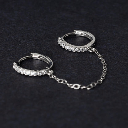 Silver huggie earrings with sparkling cubic zirconia detailing, connected by a delicate chain, displayed on a dark background.