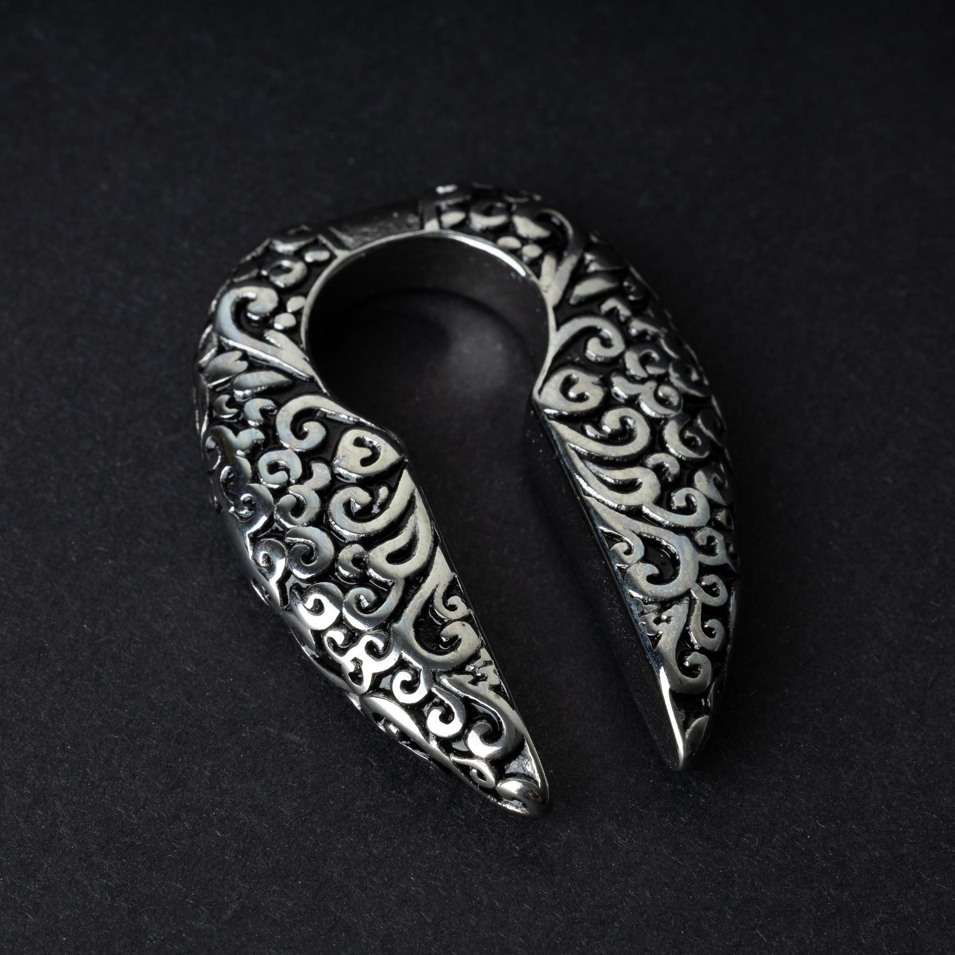 A pair of split ear weights in polished silver with detailed filigree patterns, displayed on a dark background.

