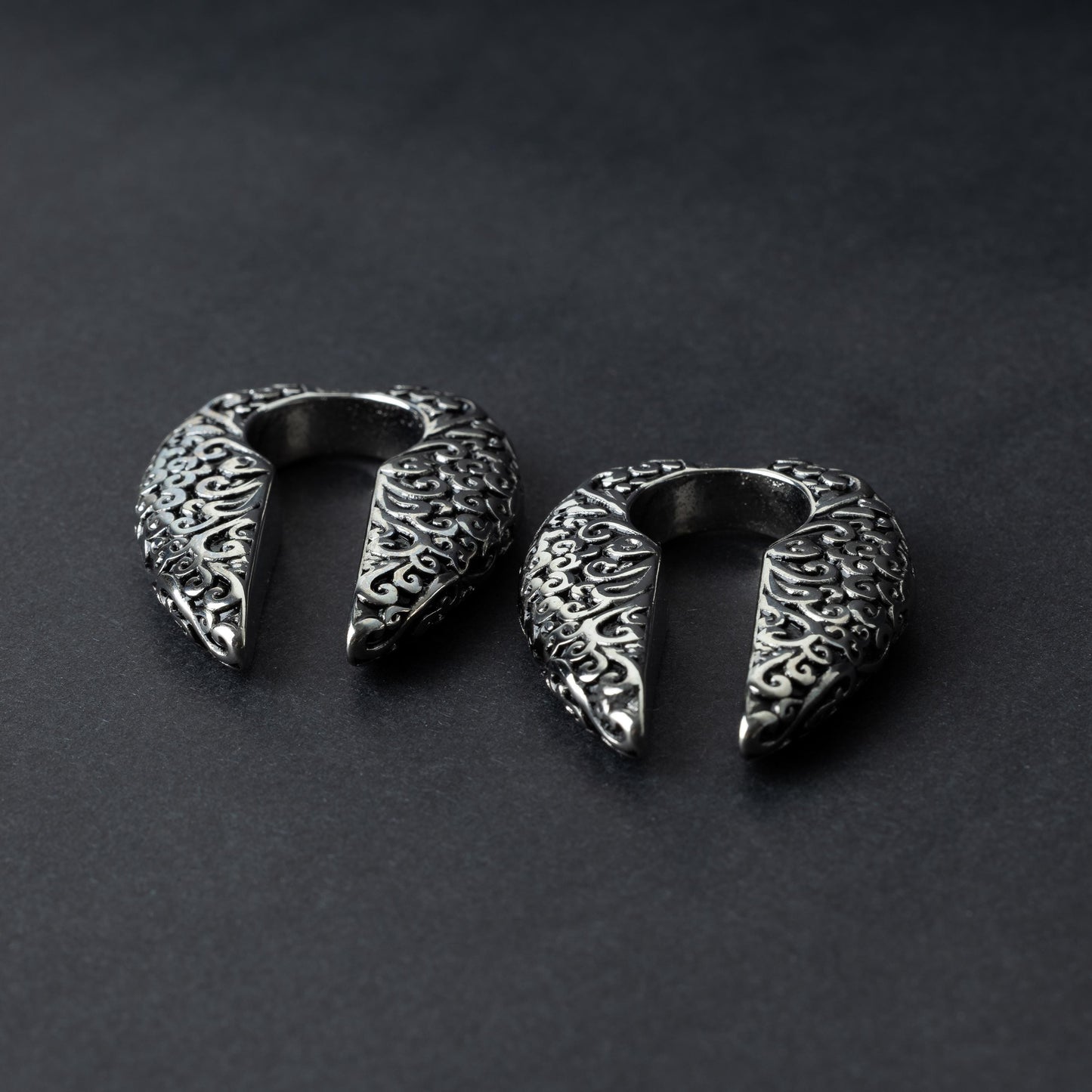 A pair of split ear weights in polished silver with detailed filigree patterns, displayed on a dark background.

