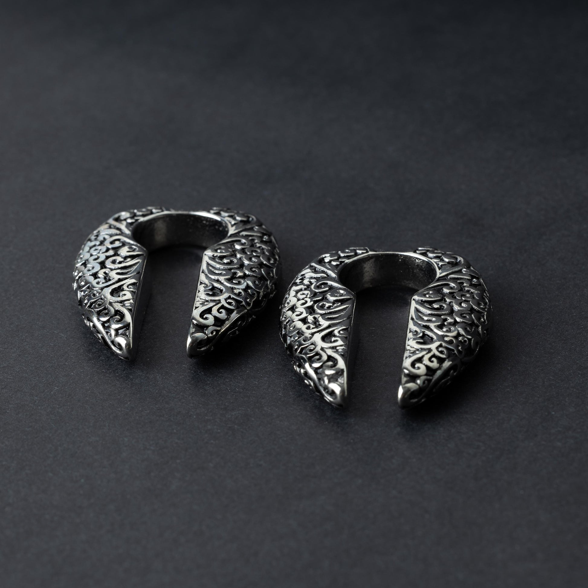 A pair of split ear weights in polished silver with detailed filigree patterns, displayed on a dark background.

