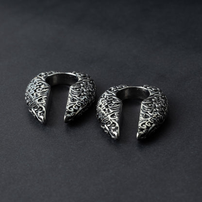 A pair of split ear weights in polished silver with detailed filigree patterns, displayed on a dark background.

