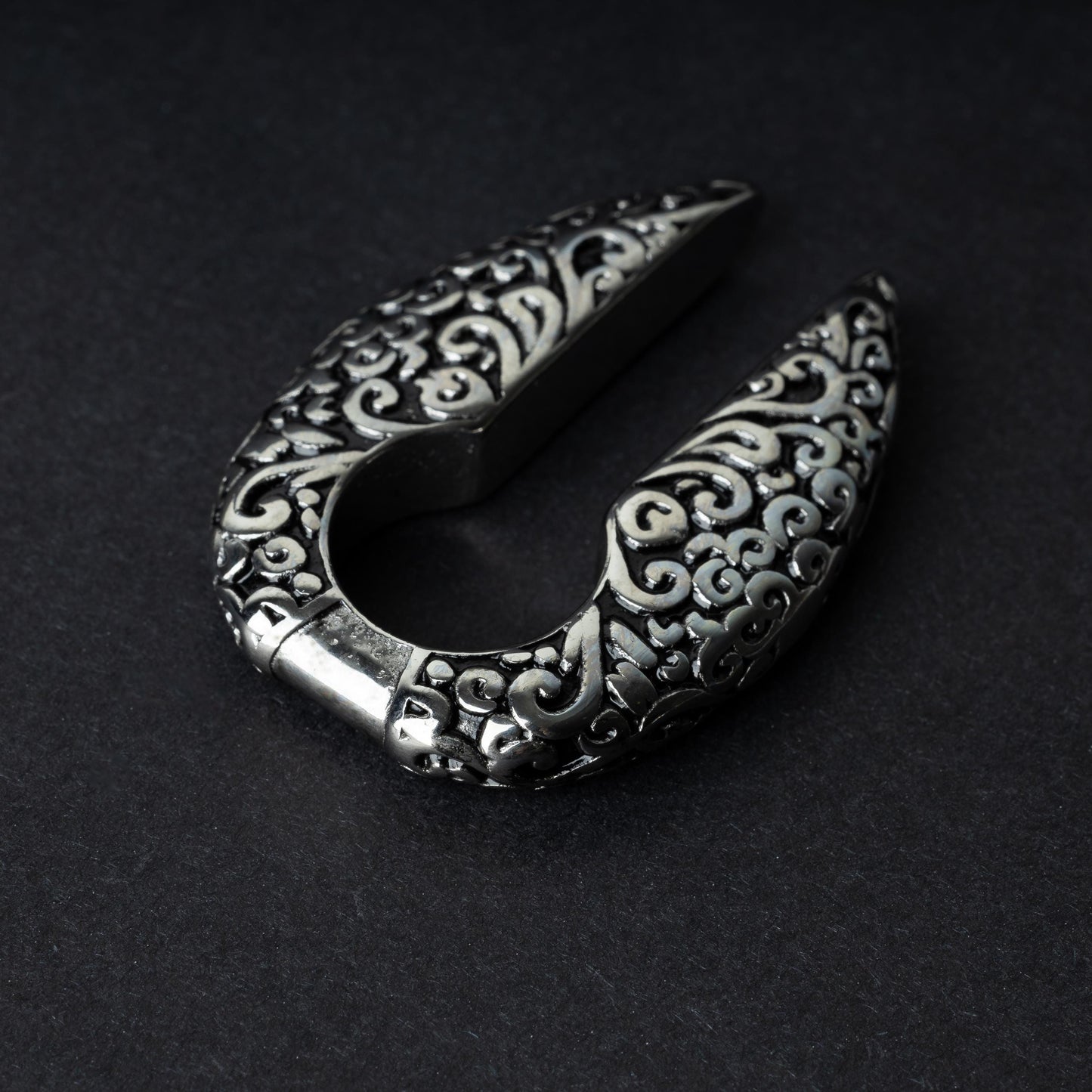 A pair of split ear weights in polished silver with detailed filigree patterns, displayed on a dark background.


