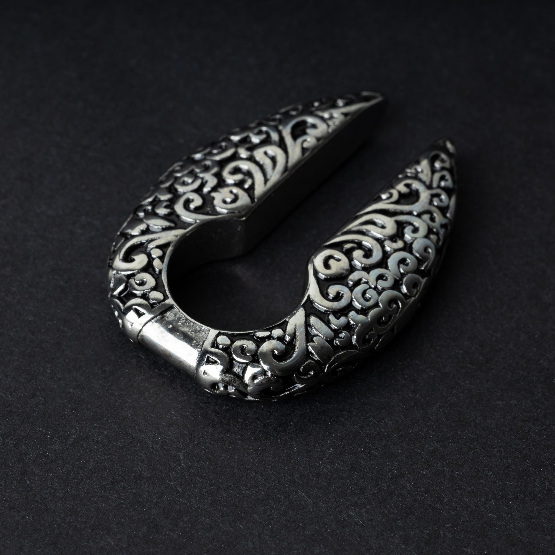 A pair of split ear weights in polished silver with detailed filigree patterns, displayed on a dark background.

