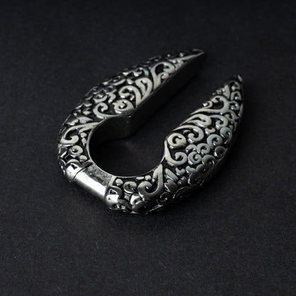 A pair of split ear weights in polished silver with detailed filigree patterns, displayed on a dark background.

