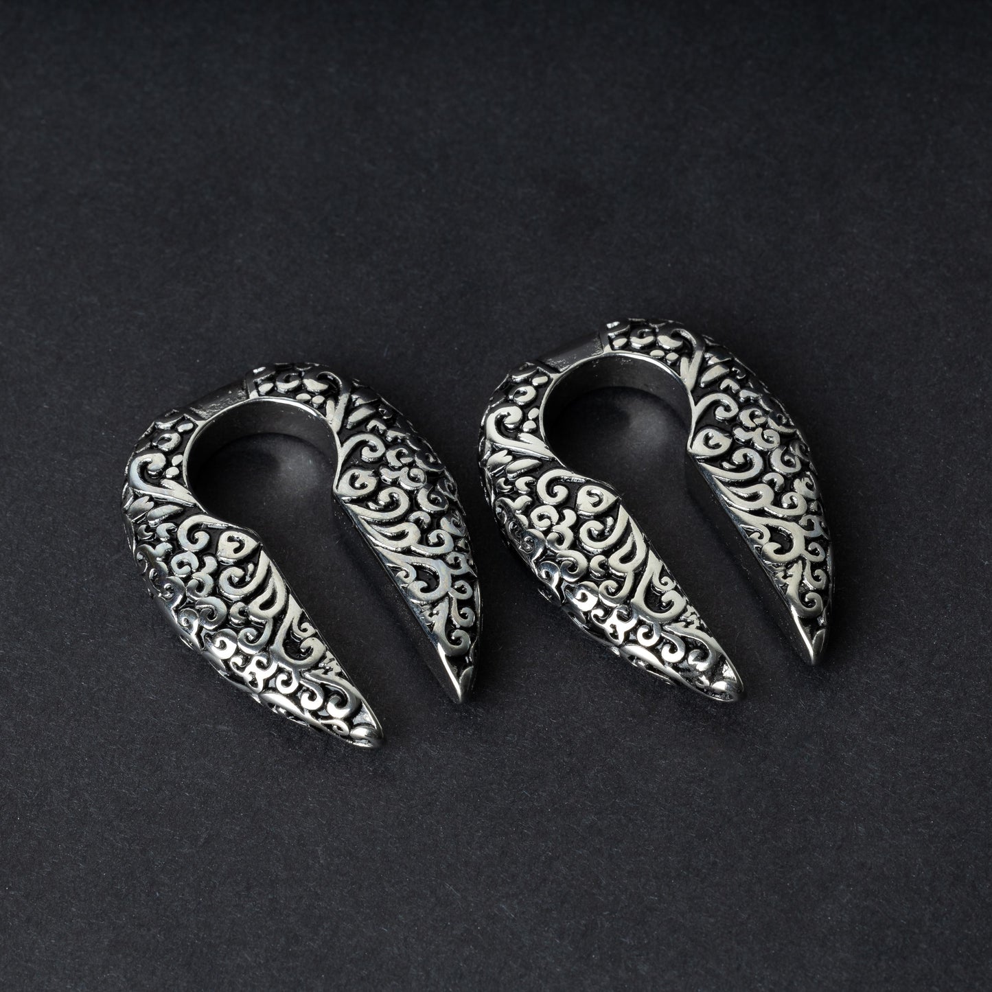 A pair of split ear weights in polished silver with detailed filigree patterns, displayed on a dark background.

