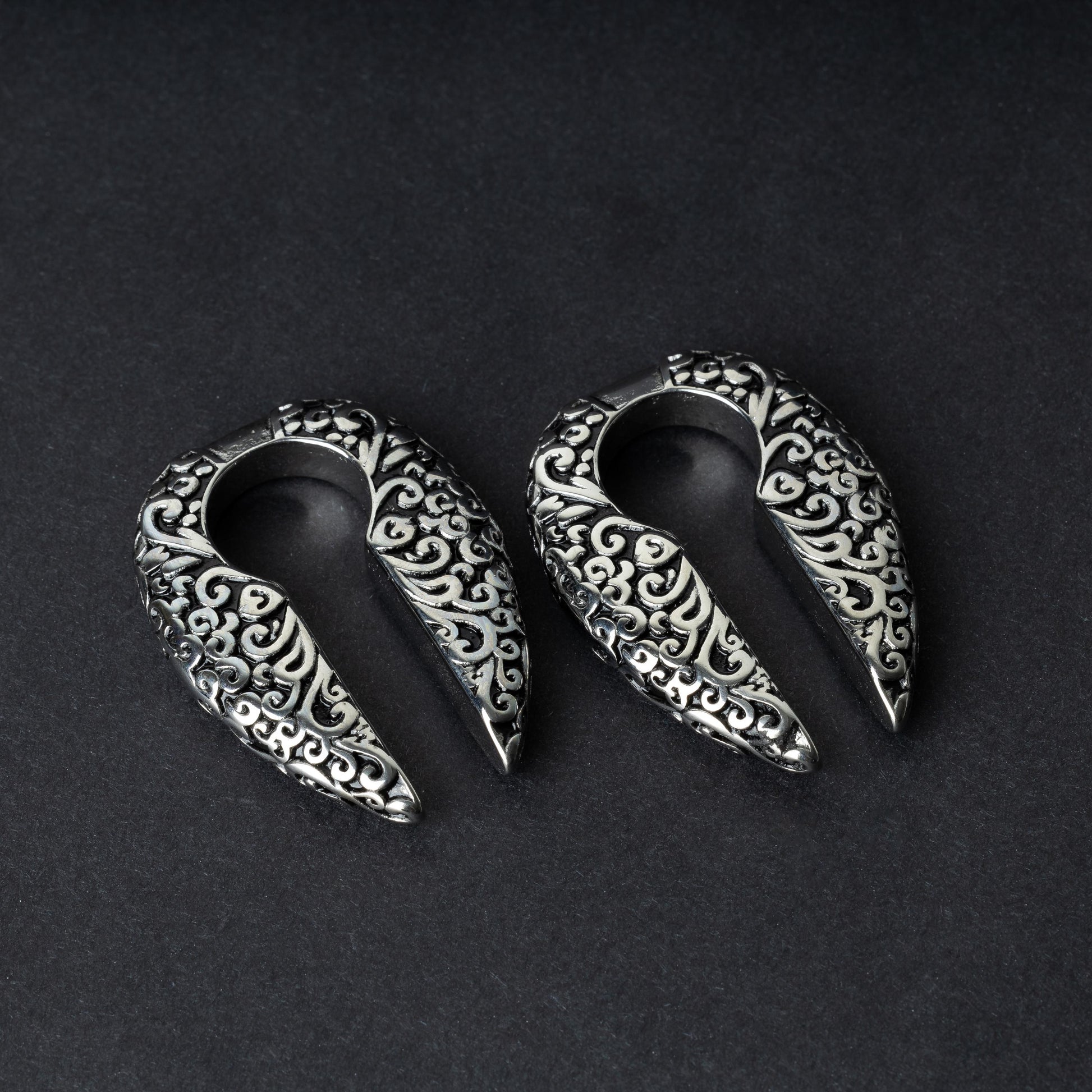 A pair of split ear weights in polished silver with detailed filigree patterns, displayed on a dark background.

