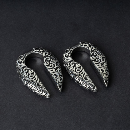 A pair of split ear weights in polished silver with detailed filigree patterns, displayed on a dark background.

