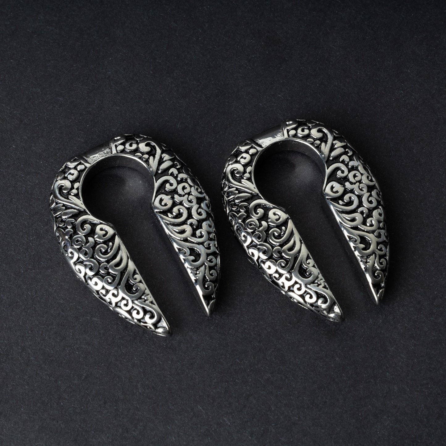A pair of split ear weights in polished silver with detailed filigree patterns, displayed on a dark background.


