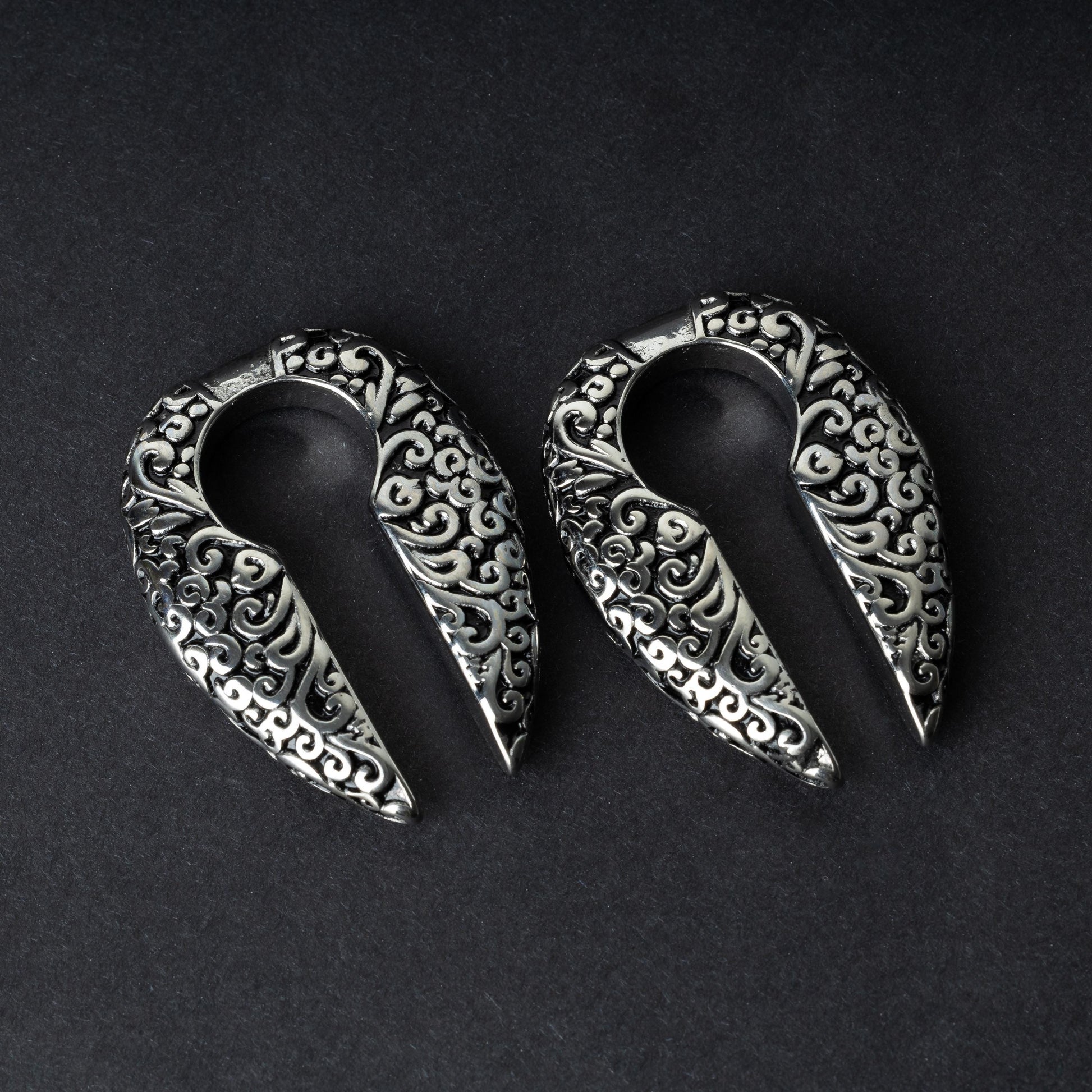 A pair of split ear weights in polished silver with detailed filigree patterns, displayed on a dark background.

