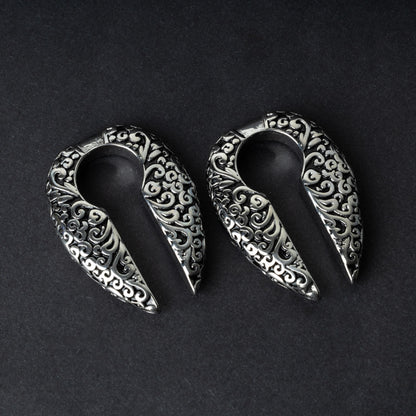 A pair of split ear weights in polished silver with detailed filigree patterns, displayed on a dark background.

