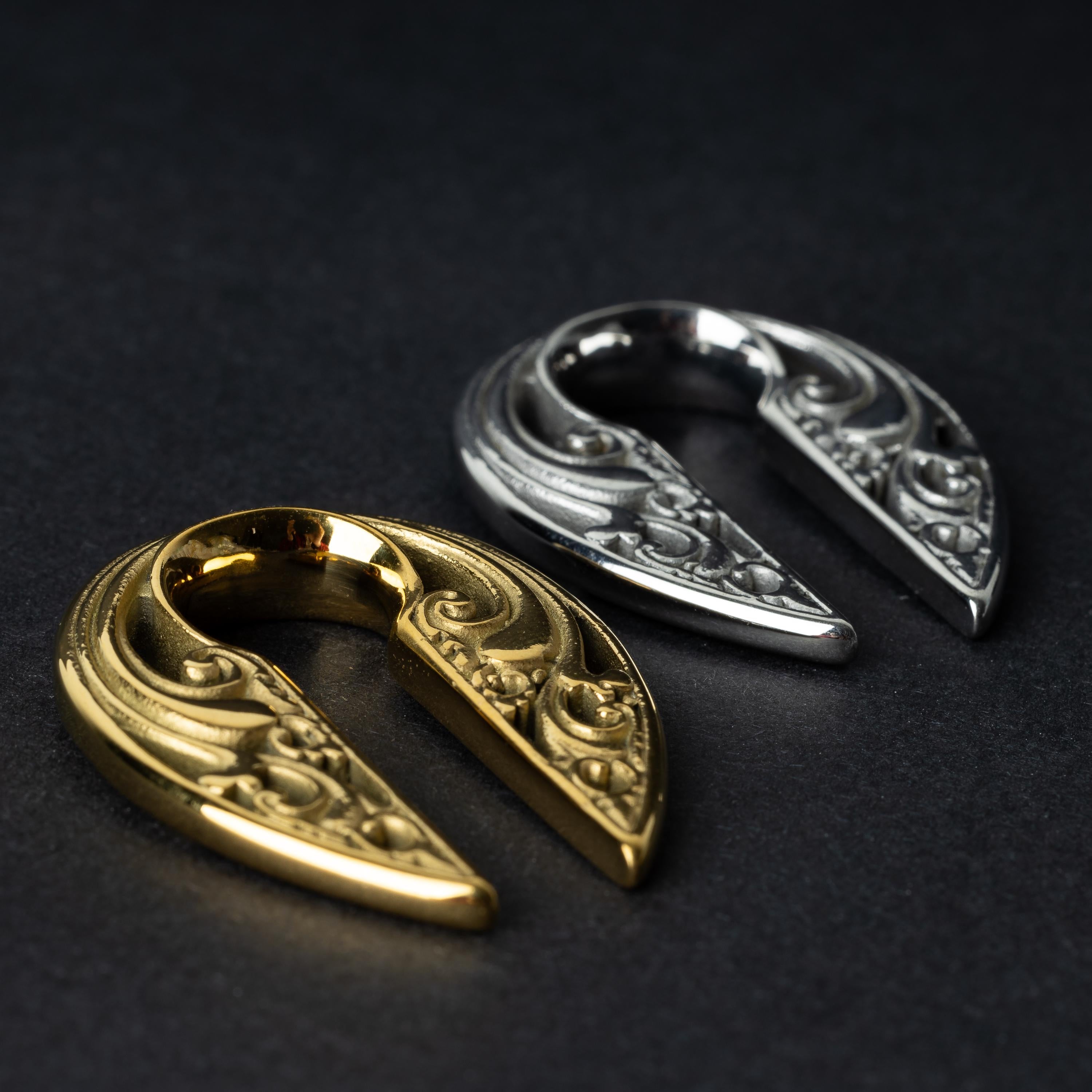 Two split ear weights with intricate filigree detailing, one in polished gold and the other in polished silver, displayed on a dark background.

