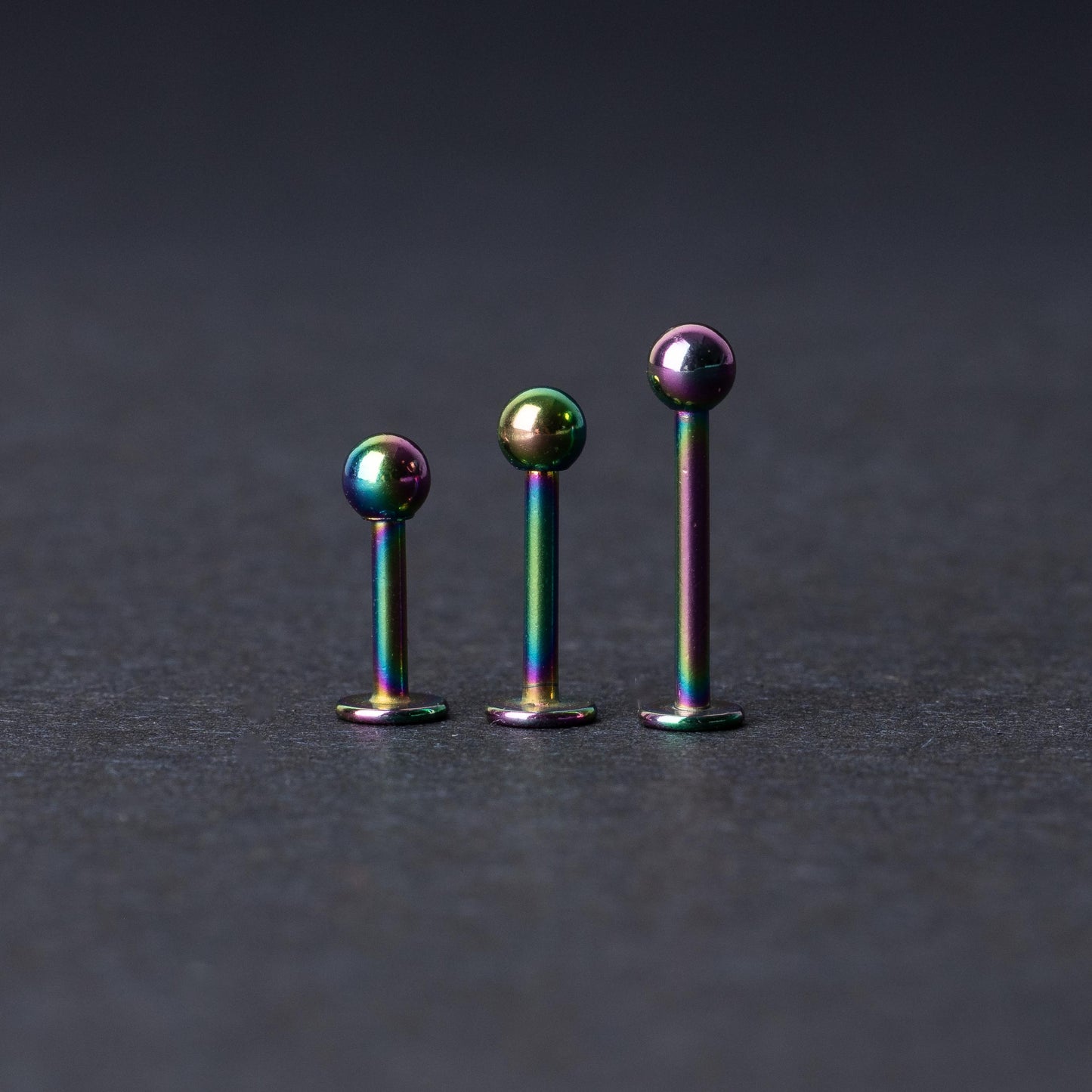 Three titanium labret studs with a rainbow iridescent finish, displayed upright on a dark textured background