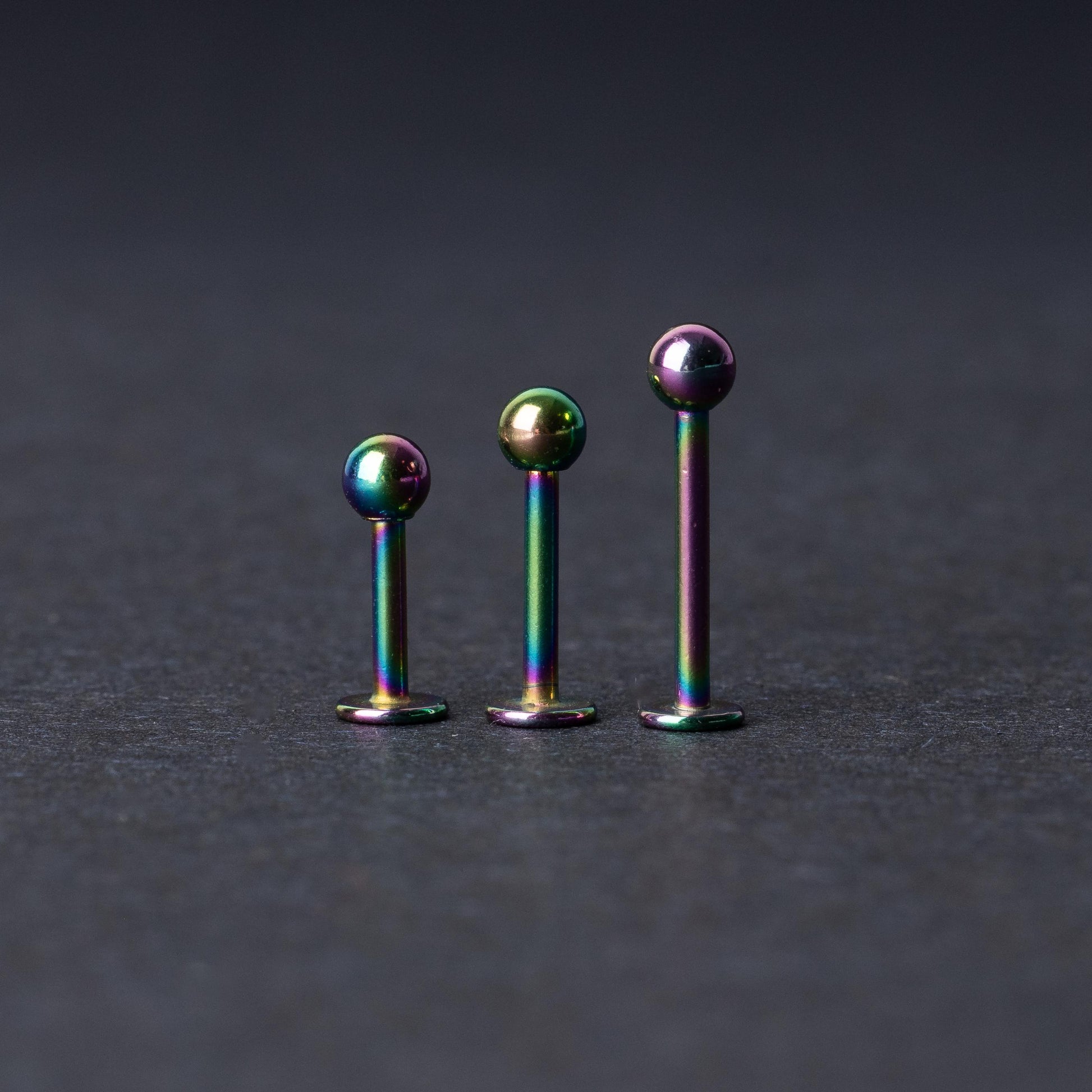 Three titanium labret studs with a rainbow iridescent finish, displayed upright on a dark textured background
