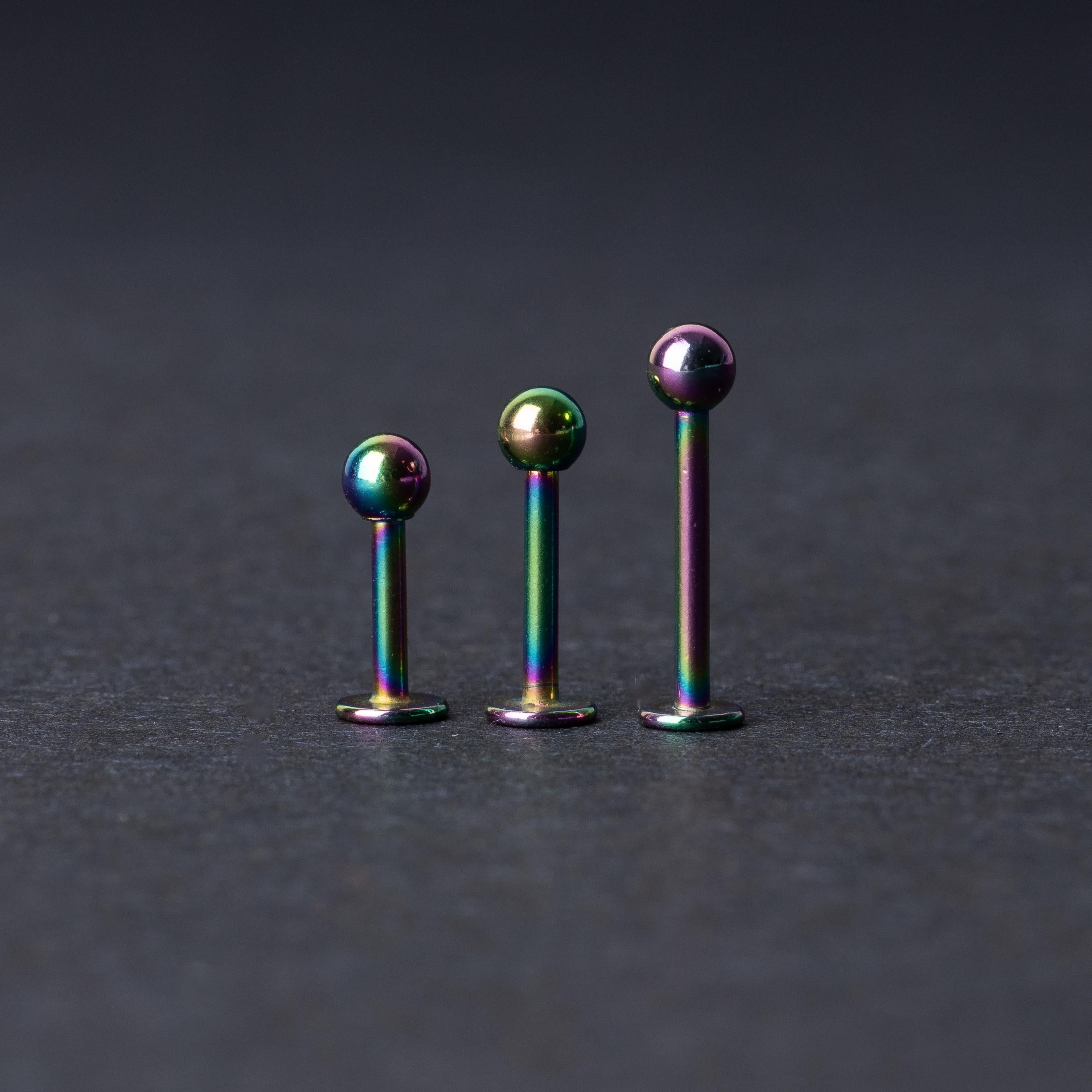 Three titanium labret studs with a rainbow iridescent finish, displayed upright on a dark textured background
