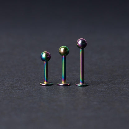 Three titanium labret studs with a rainbow iridescent finish, displayed upright on a dark textured background