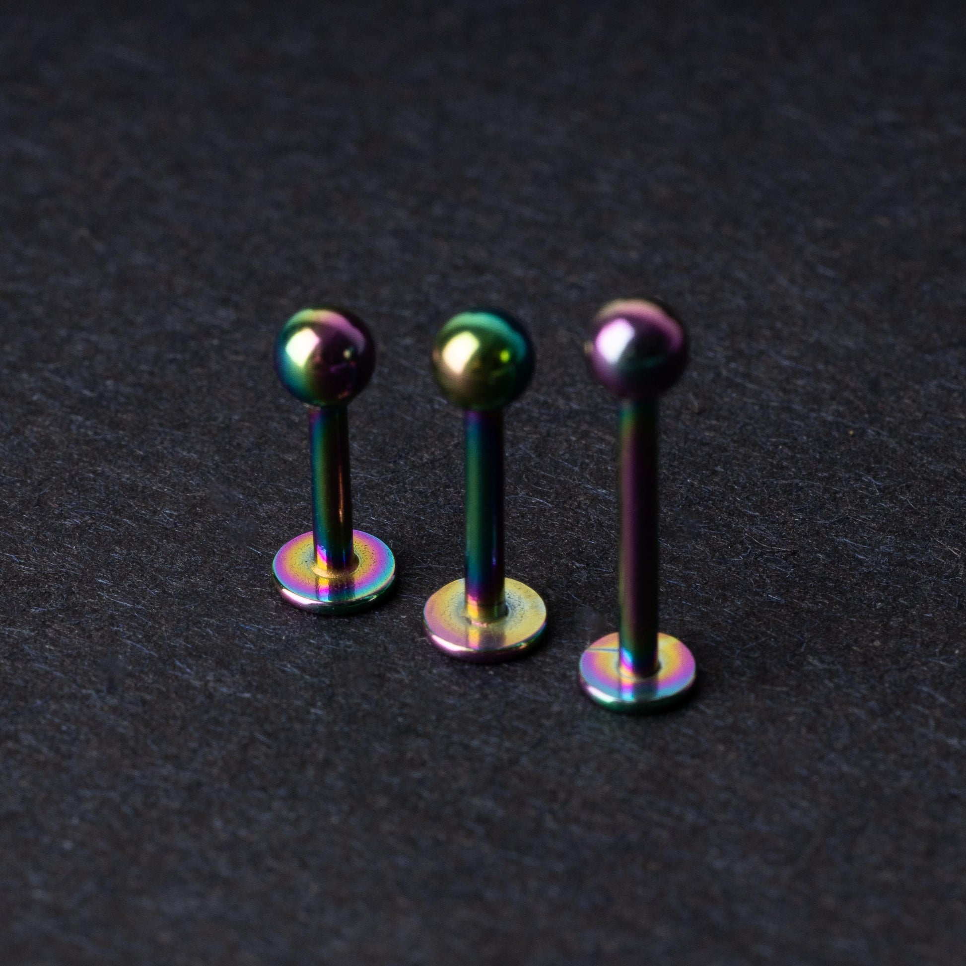 Three titanium labret studs with a rainbow iridescent finish, displayed upright on a dark textured background