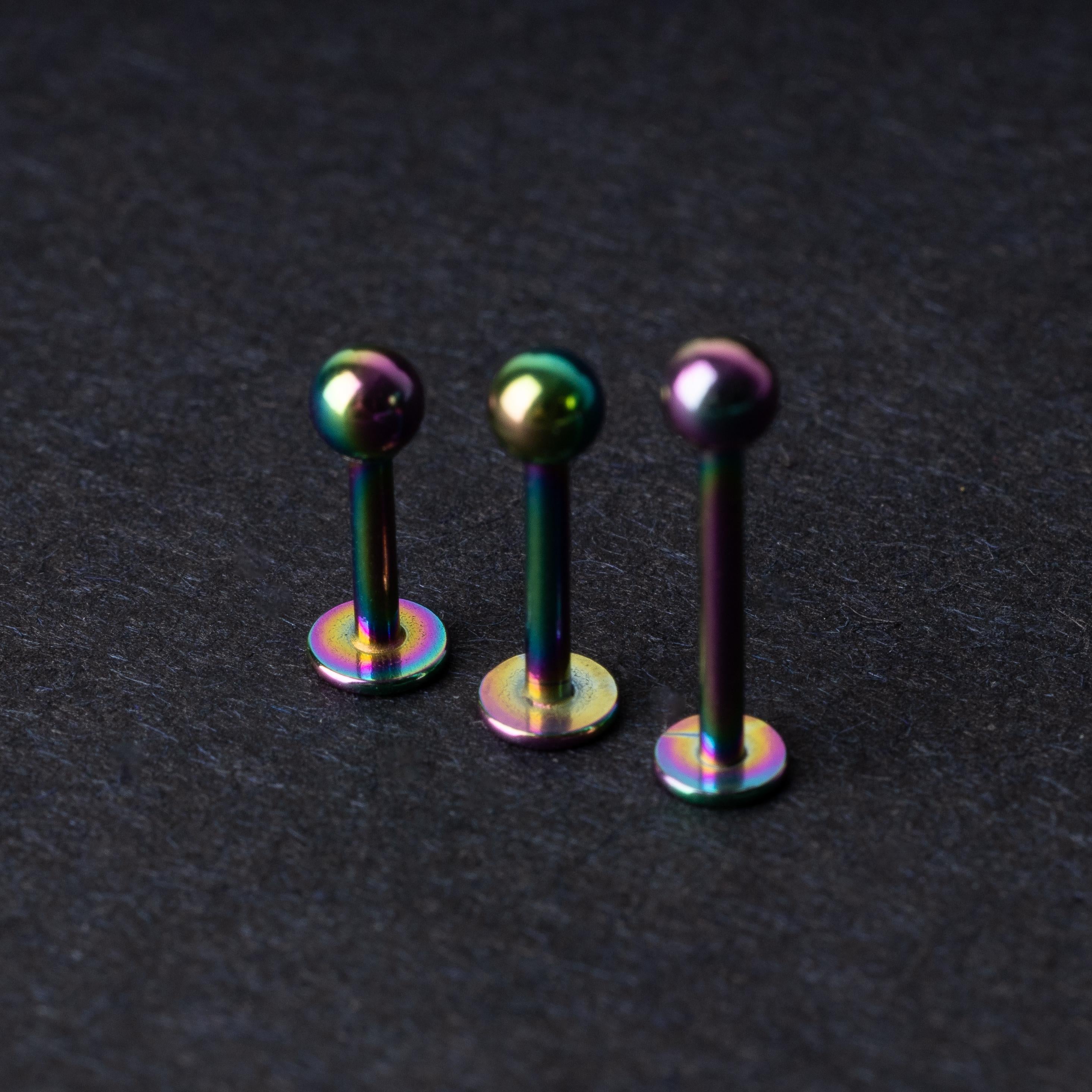 Three titanium labret studs with a rainbow iridescent finish, displayed upright on a dark textured background