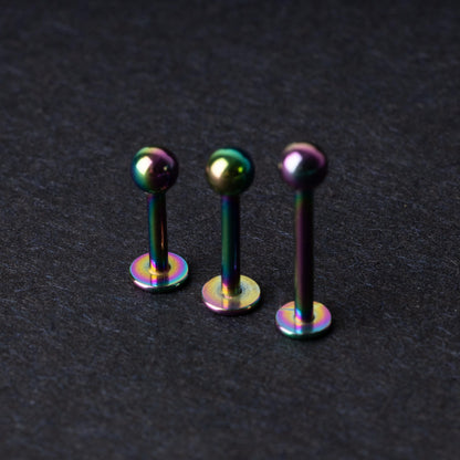 Three titanium labret studs with a rainbow iridescent finish, displayed upright on a dark textured background