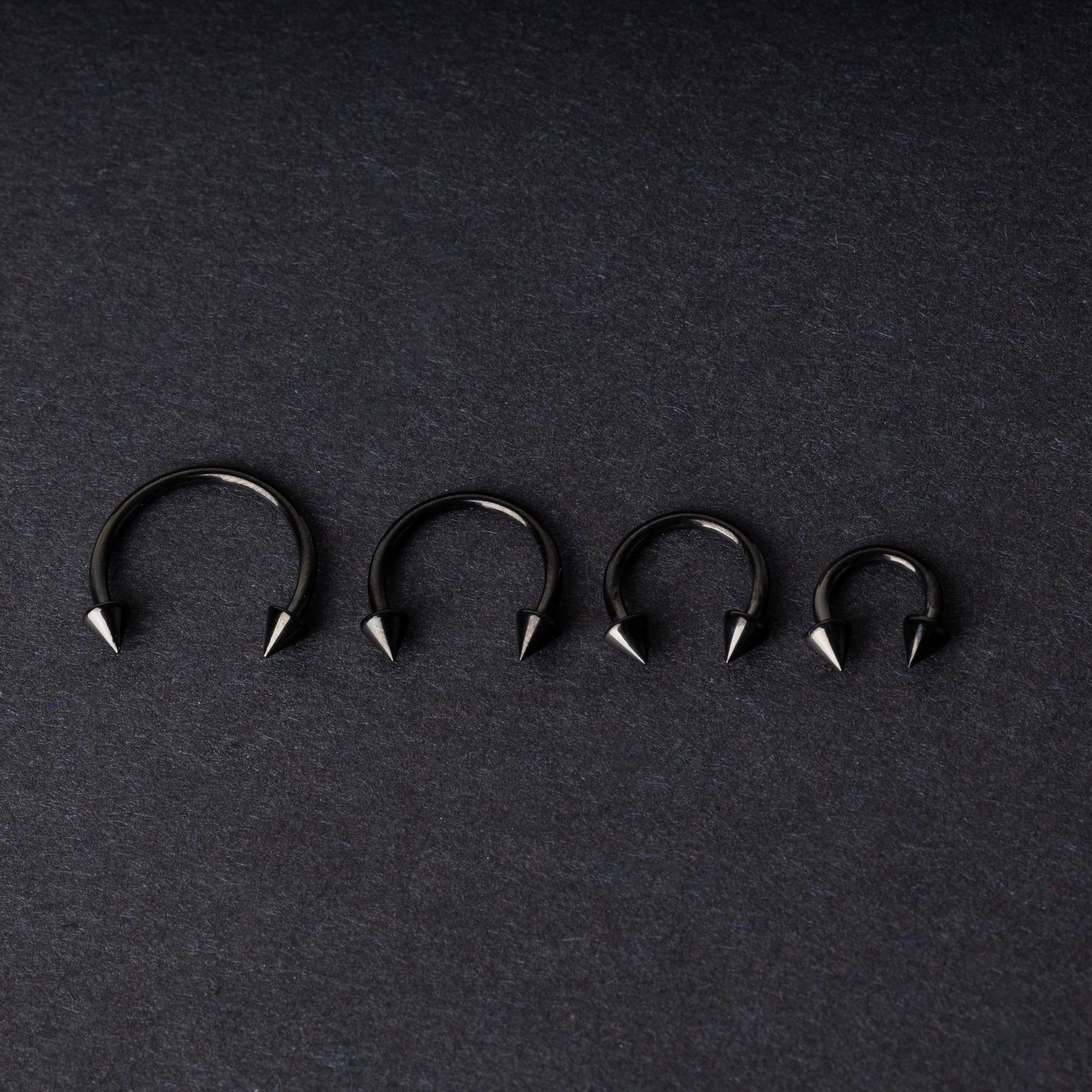 Four G23 titanium black horseshoe rings with spiked ends in varying sizes, displayed on a dark background showcasing their polished finish.