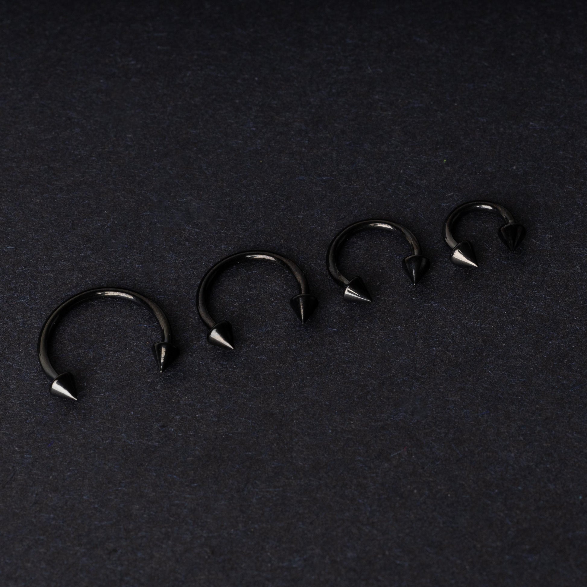 Four G23 titanium black horseshoe rings with spiked ends in varying sizes, displayed on a dark background showcasing their polished finish.
