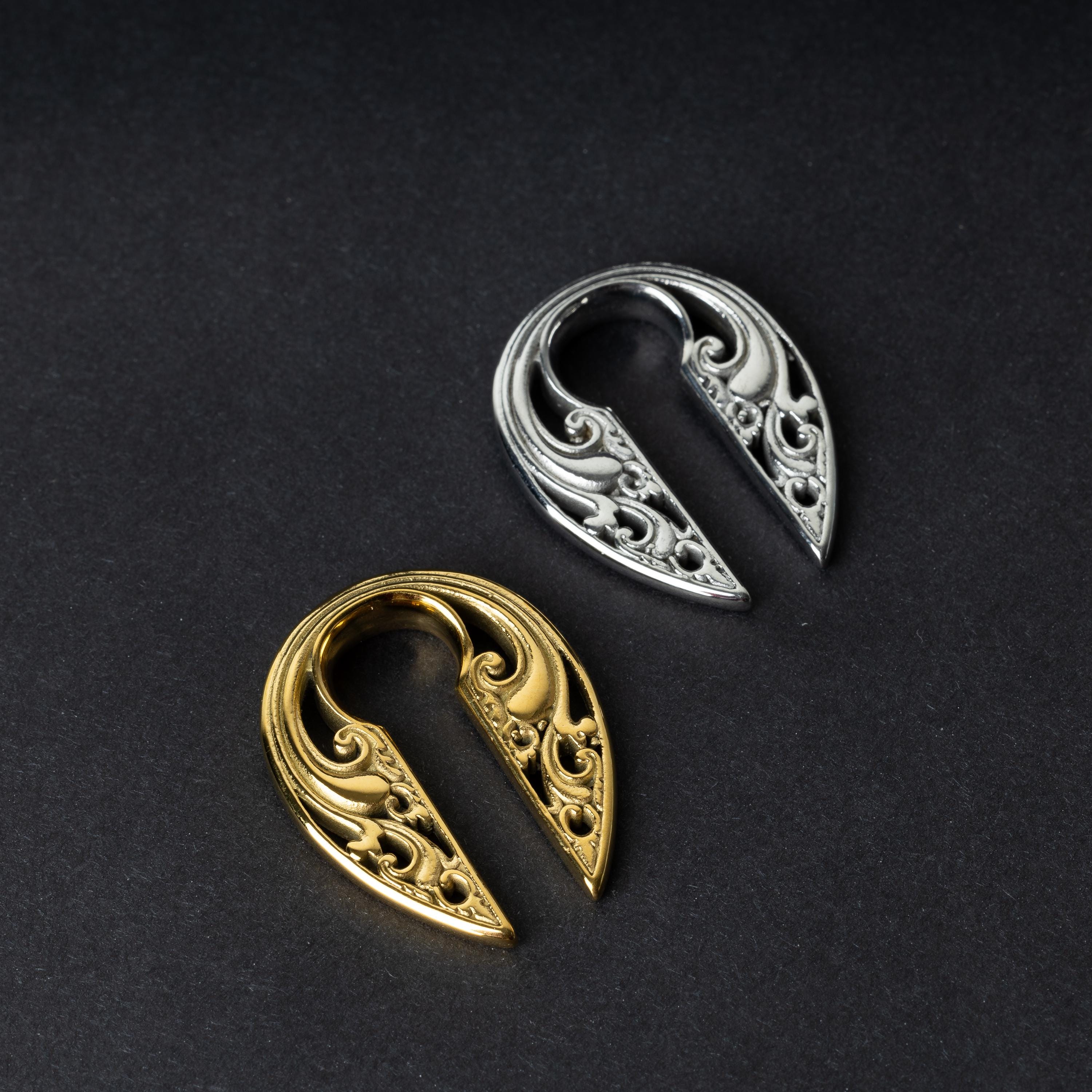 Two split ear weights with intricate filigree detailing, one in polished gold and the other in polished silver, displayed on a dark background.

