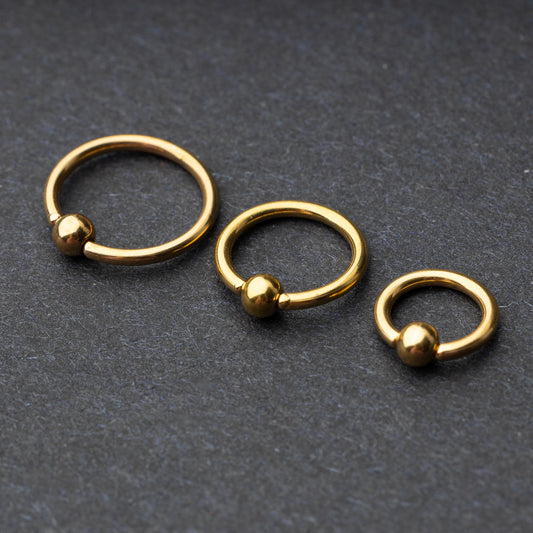 Three gold-tone stainless steel captive bead rings of varying sizes displayed on a dark background.