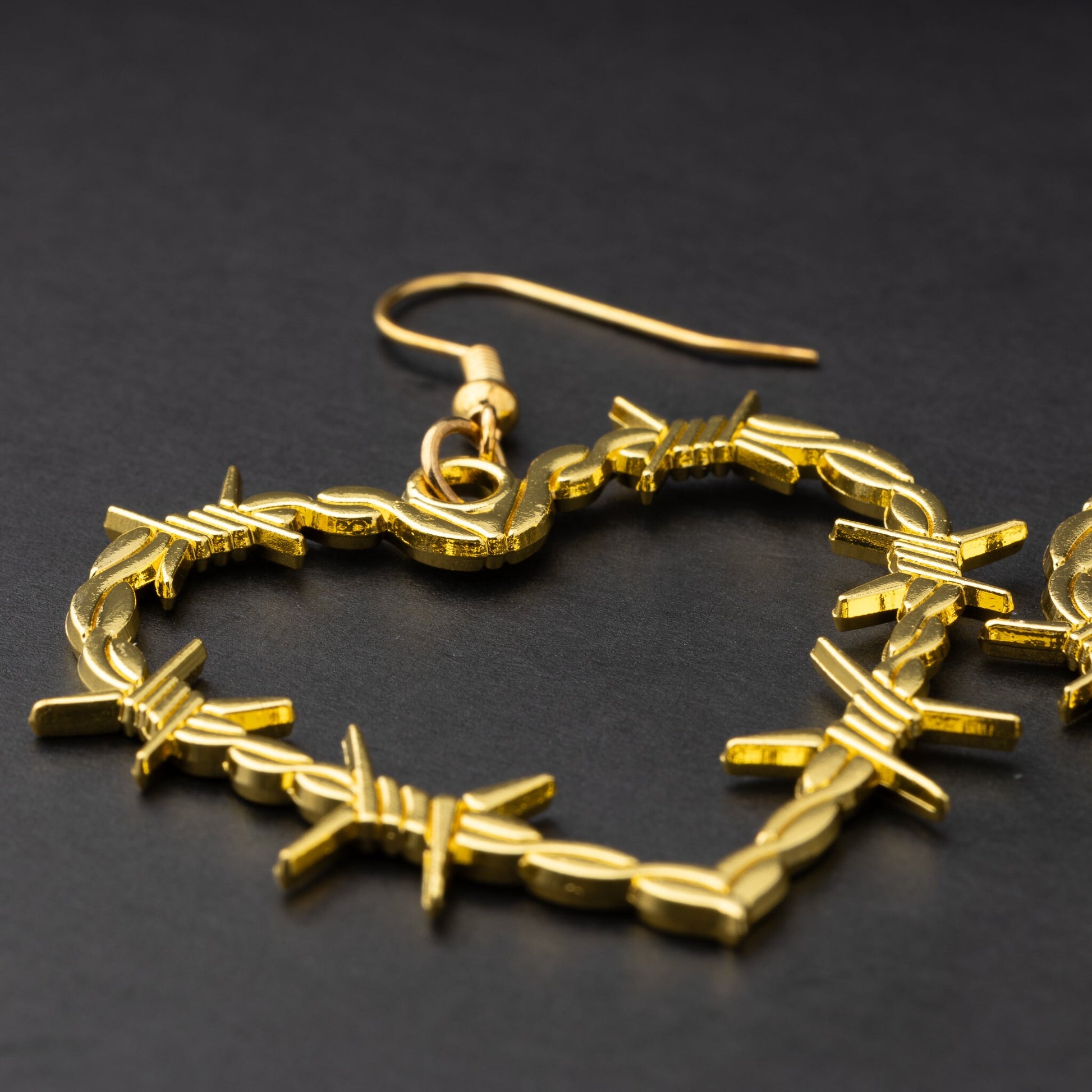 A single heart-shaped gold-tone hoop earring designed with barbed wire detailing, displayed on a dark background.