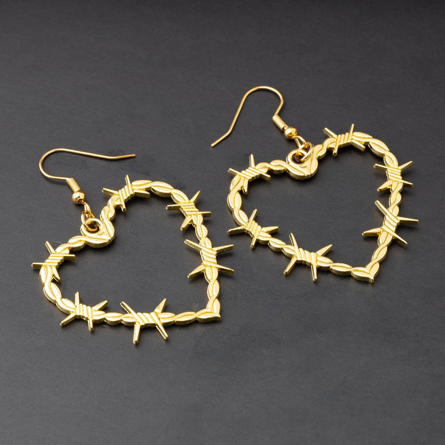 A pair of heart-shaped gold-tone hoop earrings designed with barbed wire detailing, displayed on a dark background.

