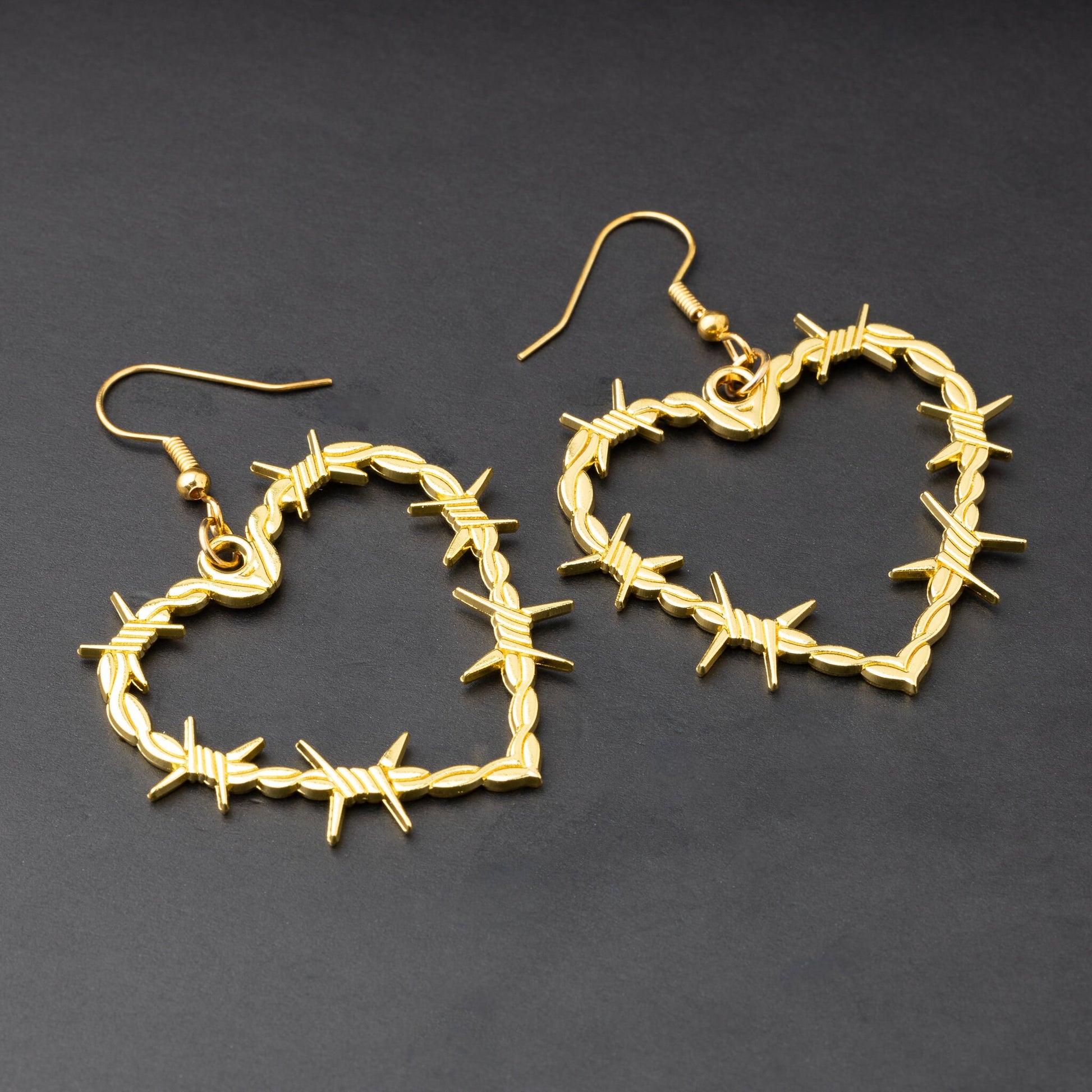A pair of heart-shaped gold-tone hoop earrings designed with barbed wire detailing, displayed on a dark background.

