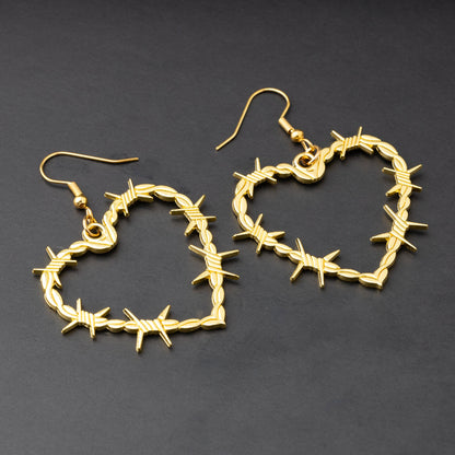 A pair of heart-shaped gold-tone hoop earrings designed with barbed wire detailing, displayed on a dark background.


