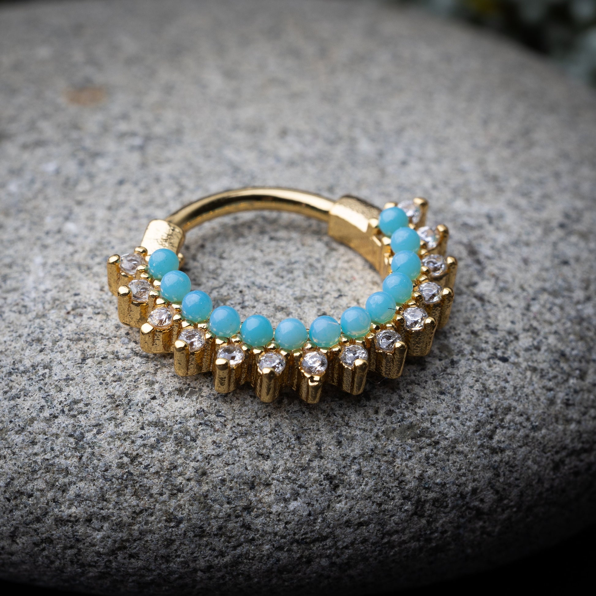 Gold-tone septum clicker with turquoise beads and cubic zirconia stones arranged in an elegant pattern, displayed on a stone background.