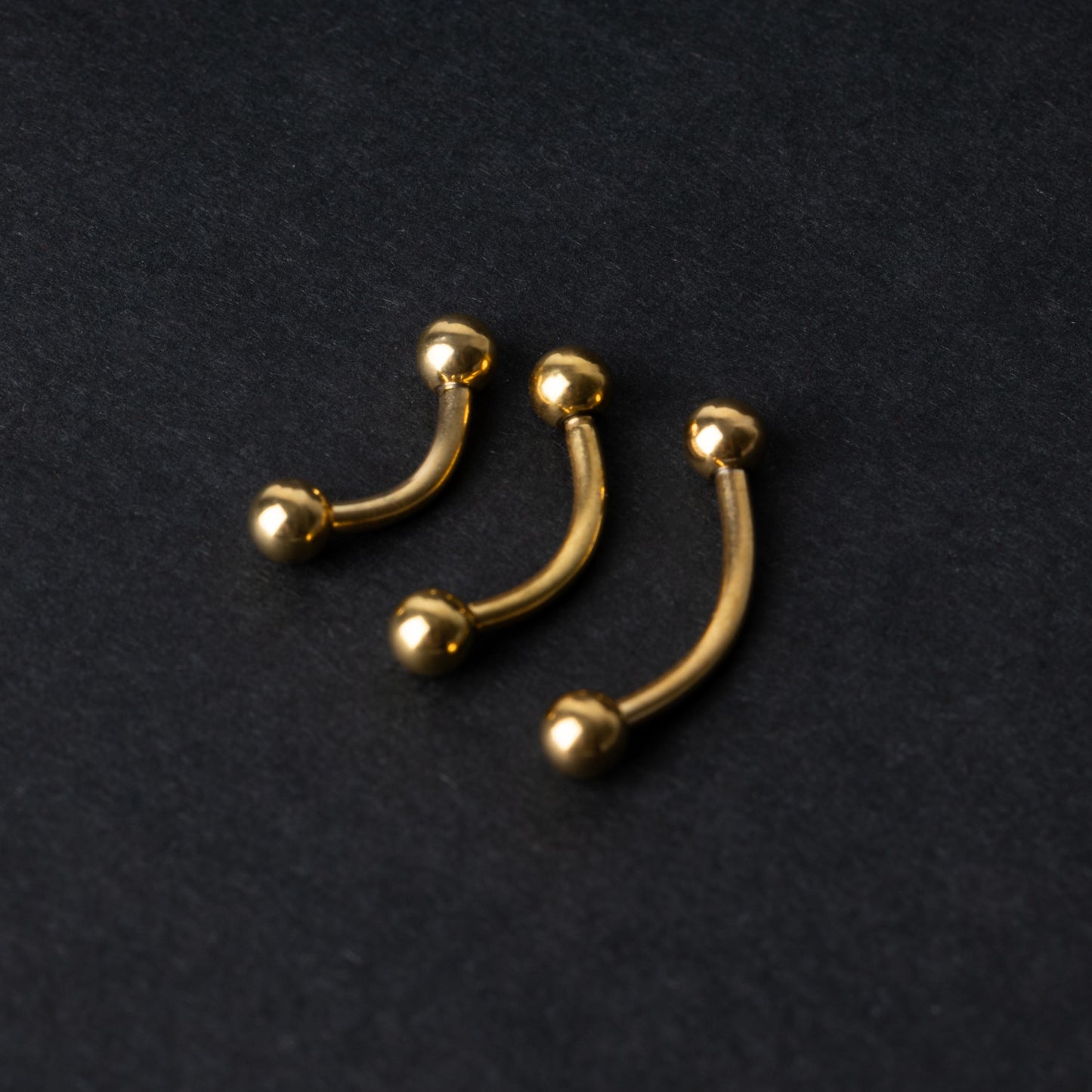 Gold curved barbells in three sizes with smooth ball ends, displayed on a dark background.
